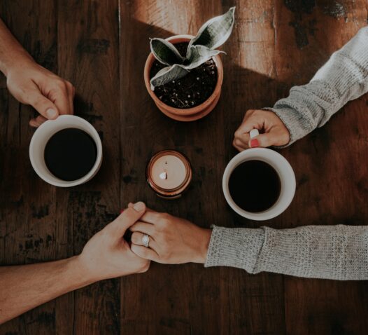 Två personer håller varandra i handen över ett bord med två kaffekoppar och en växt.