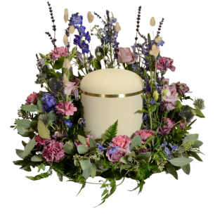 En vit urna omgiven av en krans av färgglada blommor, inklusive rosa och lila rosor samt blå och lila blommor, omgiven av gröna blad.
