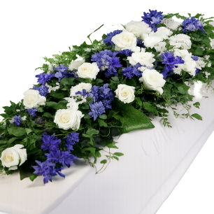 Vit kista dekorerad med vita rosor och blå blommor, omgiven av gröna blad.